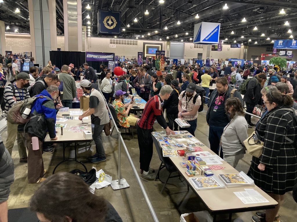 Image of crowd of people gathered around tables with board games on them. The image was taken at PAX Unplugged.
